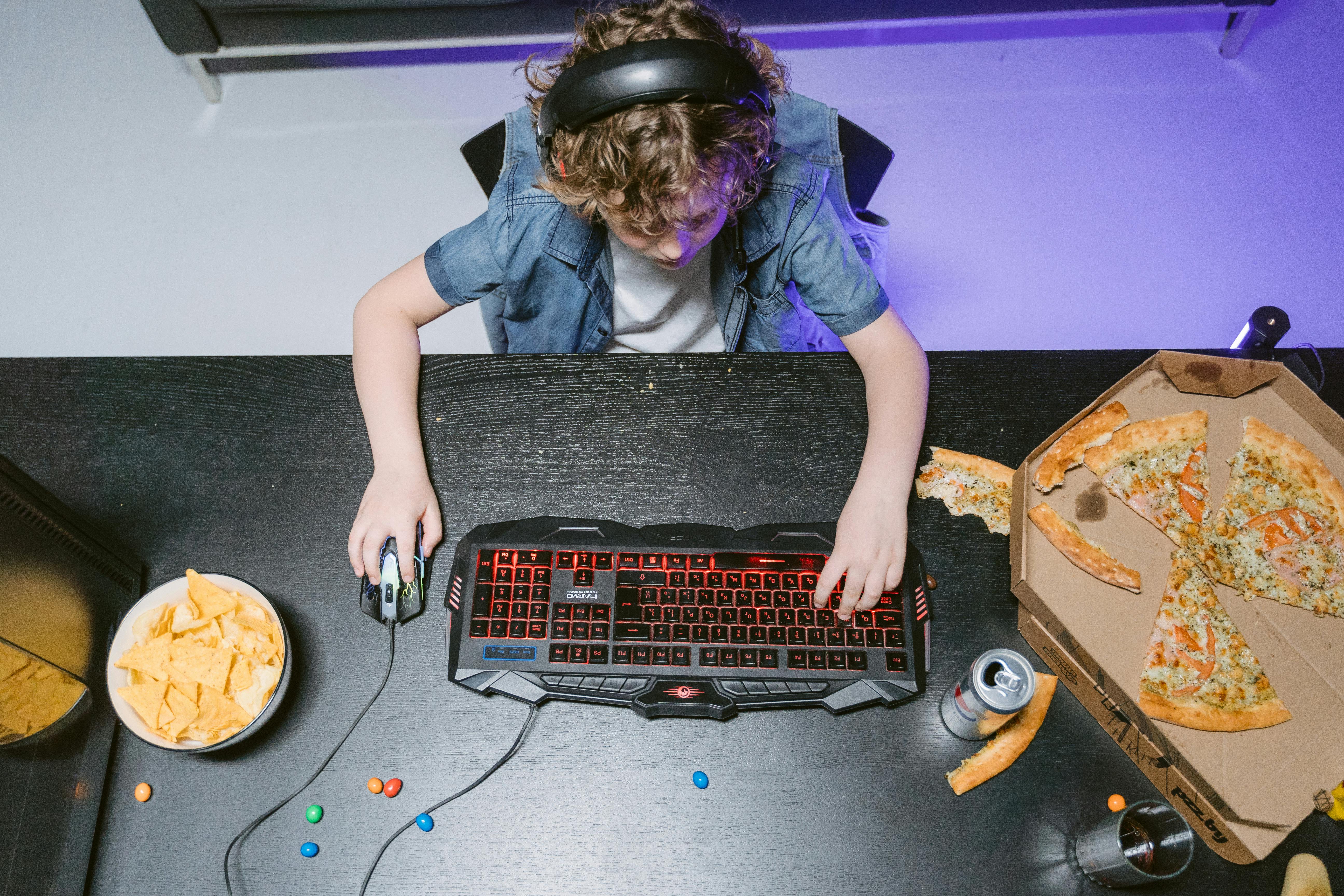 Child using a computer keyboard with pizza, chips, and candy nearby